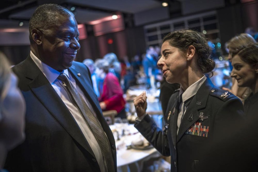 A man dressed in a business suit talks to a woman in a military uniform.
