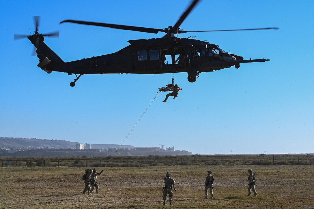 A helicopter hovers above a group of service members as a simulated casualty is hoisted upwards.