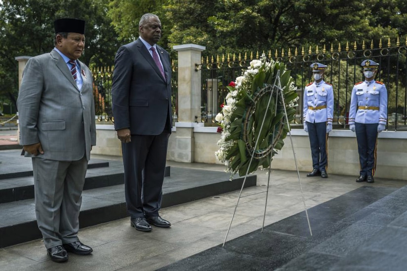 Two men in business suits look at a wreath on a stand in front of them.