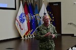 Man in military uniform stands in front of a row of flags.