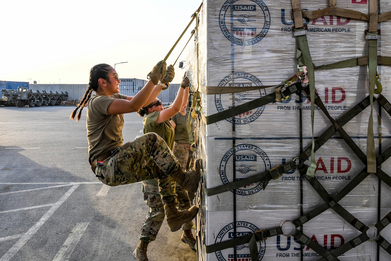 An airman pulls down on straps securing boxes onto a pallet sitting on a flightline.
