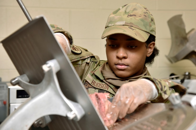 A service member works at a meat slicer.