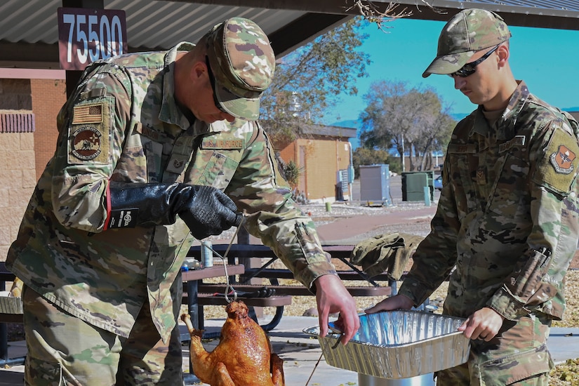 A service member holds a fried turkey upside down with a lifting hook as another service member leans forward with a roasting pan.