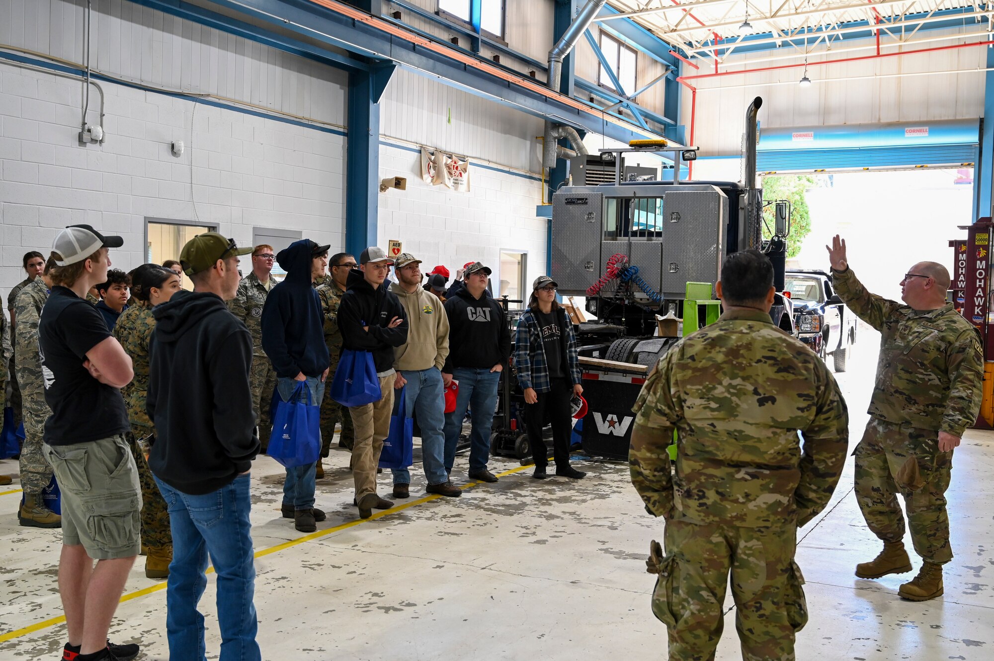 A group of civilian high school students listen to a military servicemember brief in a vehicle maintenance garage.