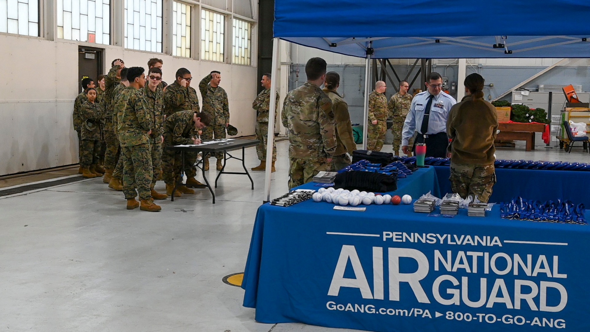 People in uniform stand in front of an Air National Guard tent inside of a hangar.