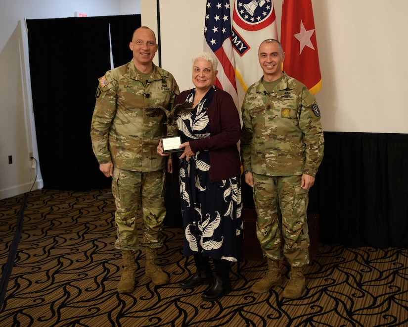 two men wearing u.s. army uniforms stand with a woman holding awards for a photo.