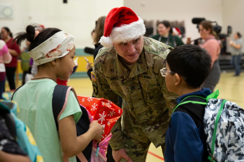 A soldier wearing a red and white Santa hat talks to two children in a gymnasium.