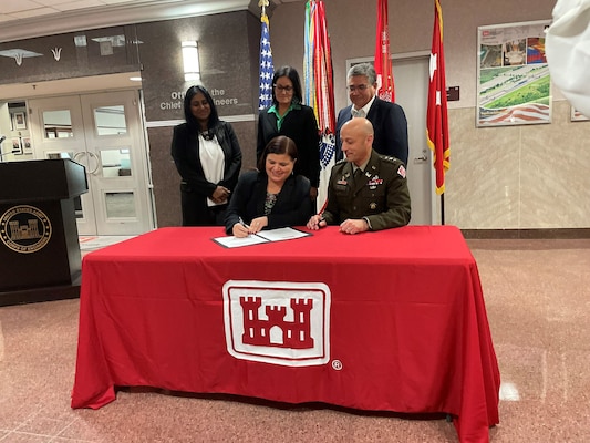 Two individuals sitting at a table signing a document as three individuals look on over their shoulders. The table is red with a U.S. Army Corps of Engineers logo on the front.