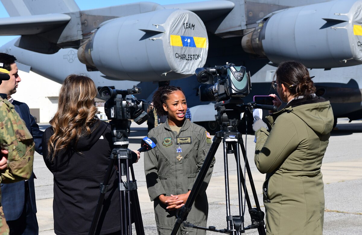 An airman stands in front of an aircraft and talks to media members.