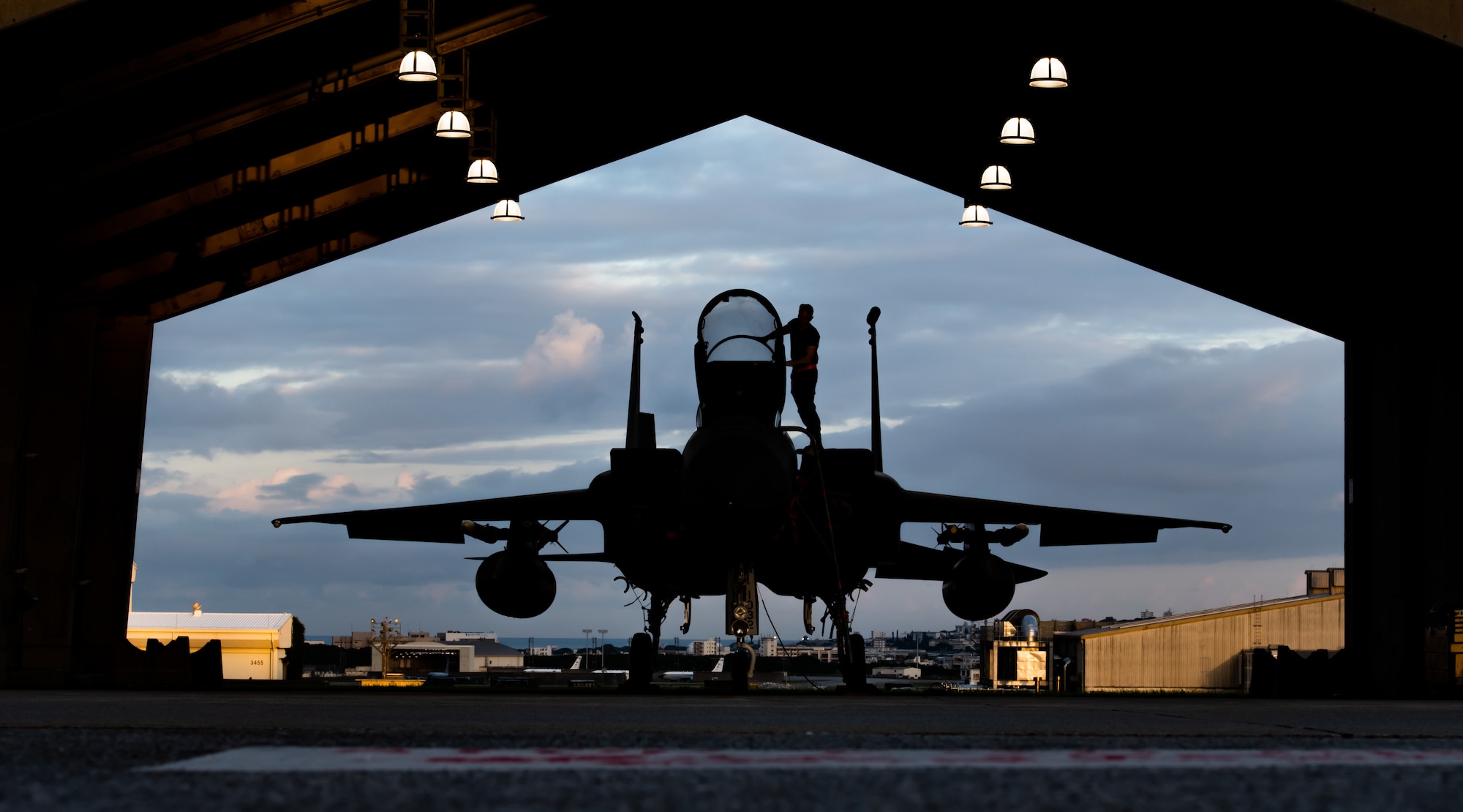 The silhouette of an Airman wiping the canopy of an F-15 centered in hangar with the sunrise in the background.