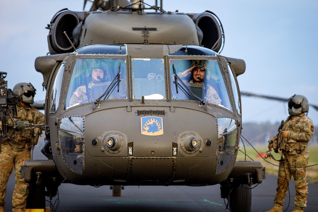 Two soldiers sit in a UH-60 Black Hawk helicopter while one salutes; two other soldiers stand outside.