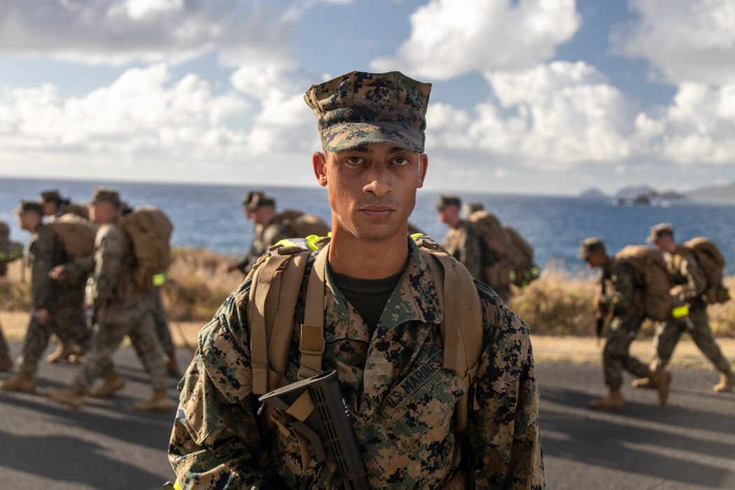A Marine in uniform staring at the camera while more Marines are walking to the left behind him.