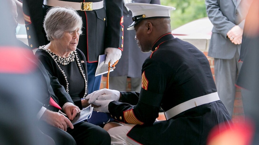 A man in dress uniform takes the hand of a seated woman.