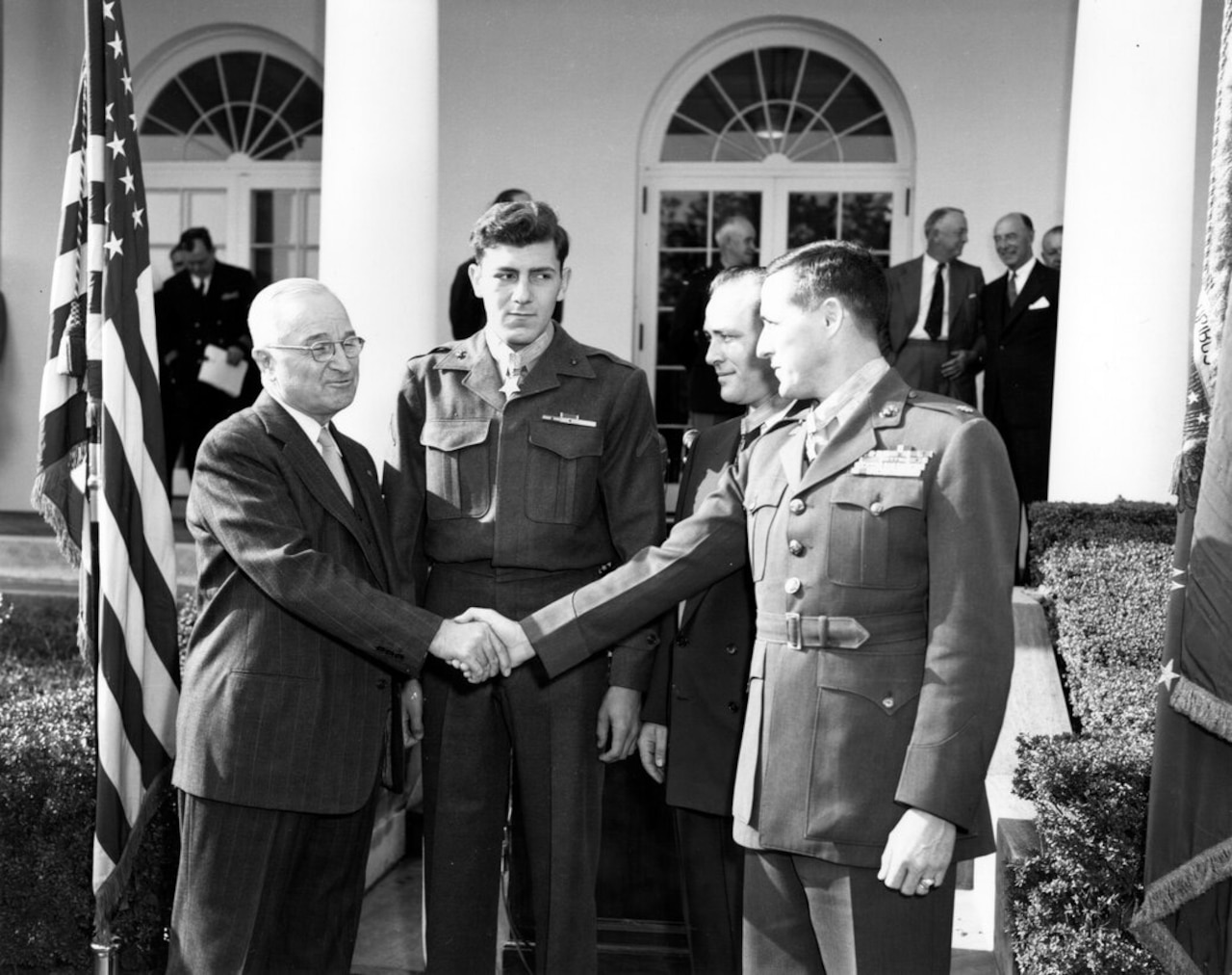 Two men shake hands as two others stand nearby. More people are in the background.