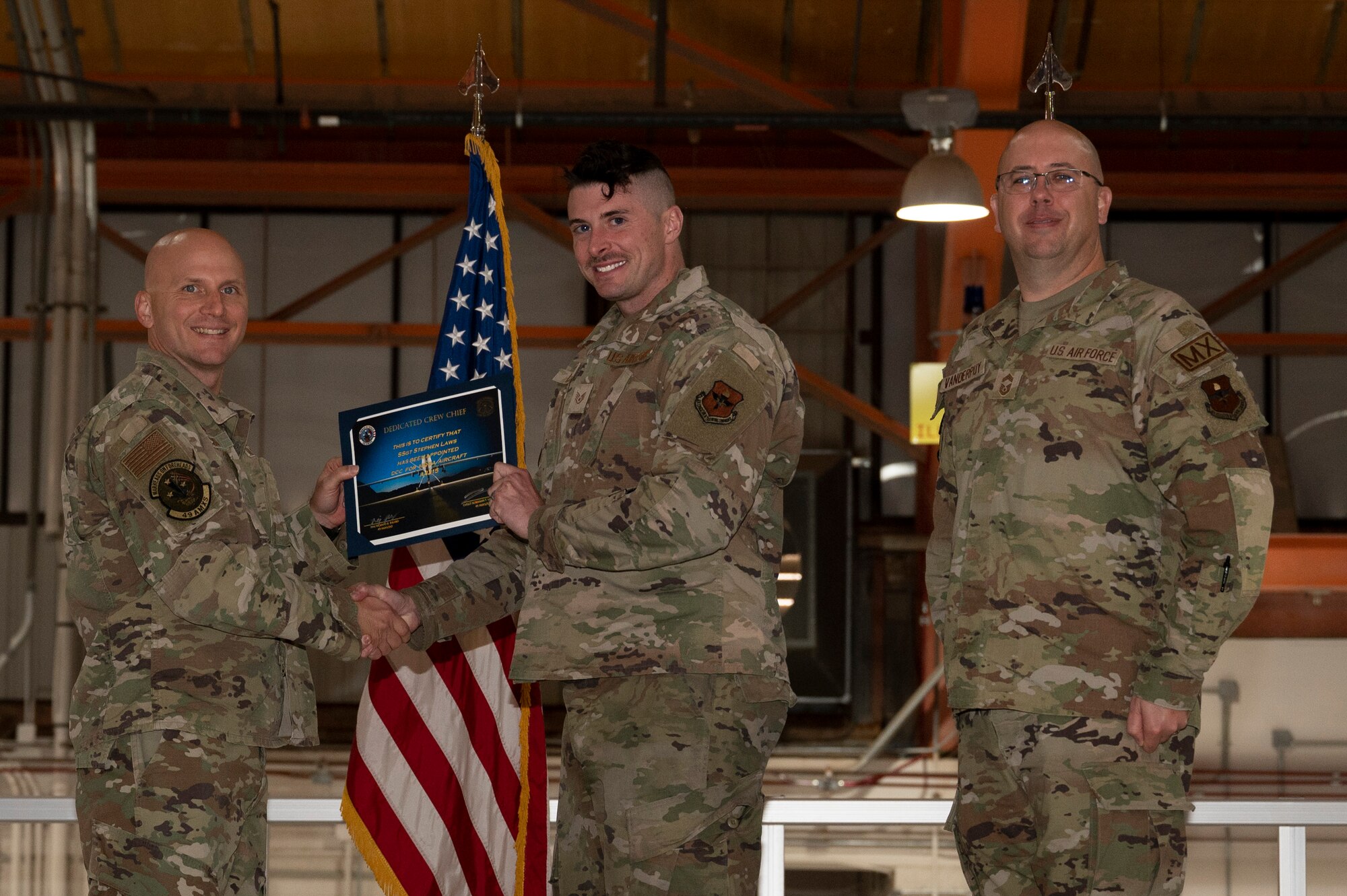 U.S. Air Force Staff Sgt. Stephen Laws, 49th Aircraft Maintenance Squadron crew chief, center, receives a certificate from U.S. Air Force Maj. Donald Bolda, 49th Aircraft Maintenance Squadron commander, left, during the 2022 Dedicated Crew Chief Appointment Ceremony at Holloman Air Force Base, New Mexico, Nov. 18, 2022. To receive the title of DCC, a crew chief must demonstrate a history of superior performance in their career, comply with all safety practices and complete all required training. (U.S. Air Force photo by Airman 1st Class Isaiah Pedrazzini)