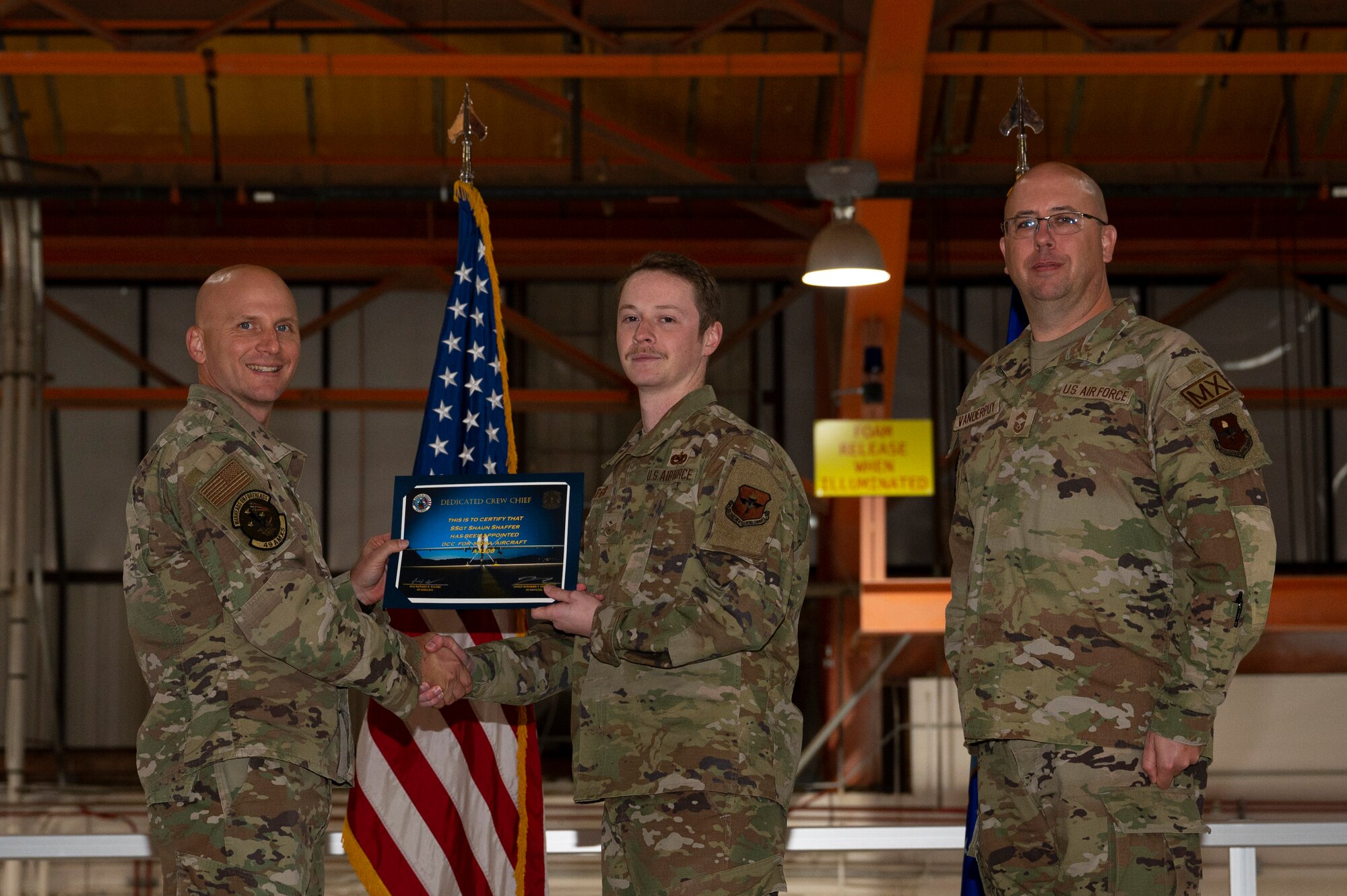 U.S. Air Force Staff Sgt. Shaun Shaffer, 49th Aircraft Maintenance Squadron crew chief, center, receives a certificate from U.S. Air Force Maj. Donald Bolda, 49th Aircraft Maintenance Squadron commander, left, during the 2022 Dedicated Crew Chief Appointment Ceremony at Holloman Air Force Base, New Mexico, Nov. 18, 2022. To receive the title of DCC, a crew chief must demonstrate a history of superior performance in their career, comply with all safety practices and complete all required training. (U.S. Air Force photo by Airman 1st Class Isaiah Pedrazzini)