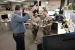 a man stands pointing and two military members stand in the background of an office