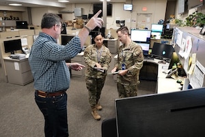 a man stands pointing and two military members stand in the background of an office