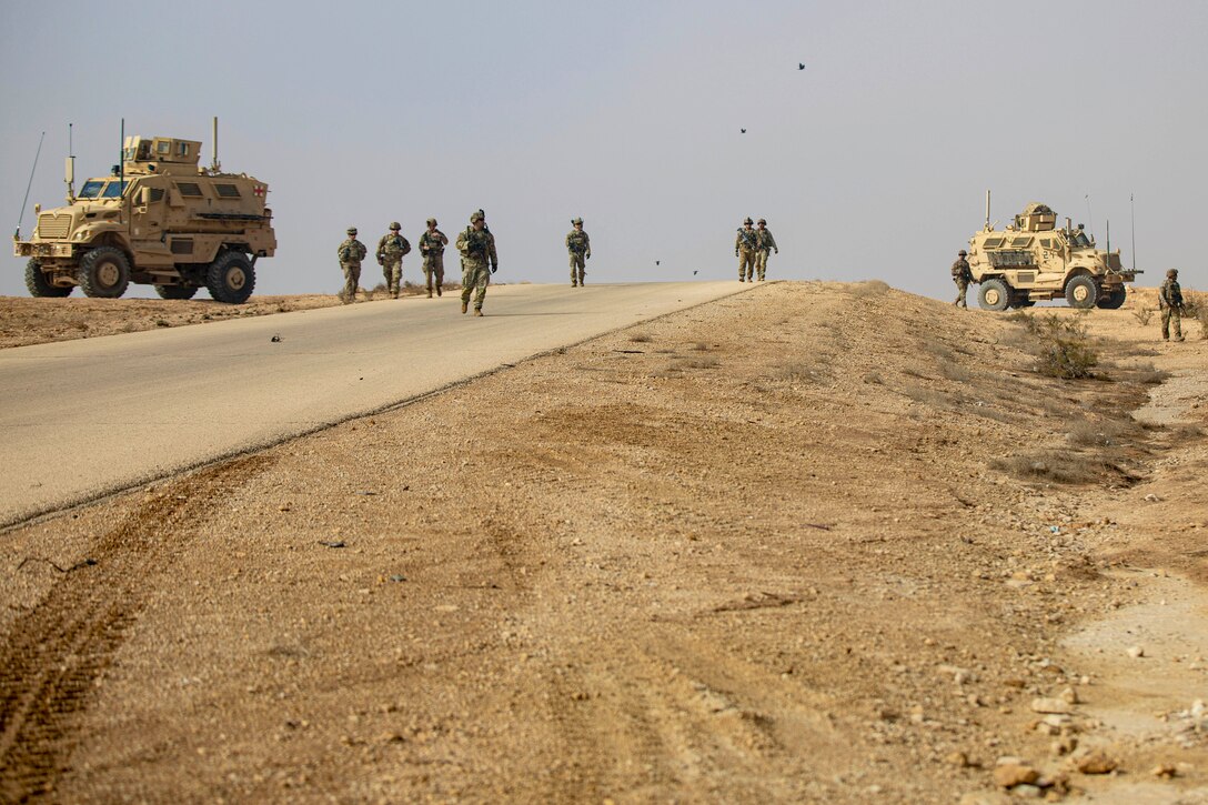 Soldiers walk on a road in a desert-like area near parked vehicles.