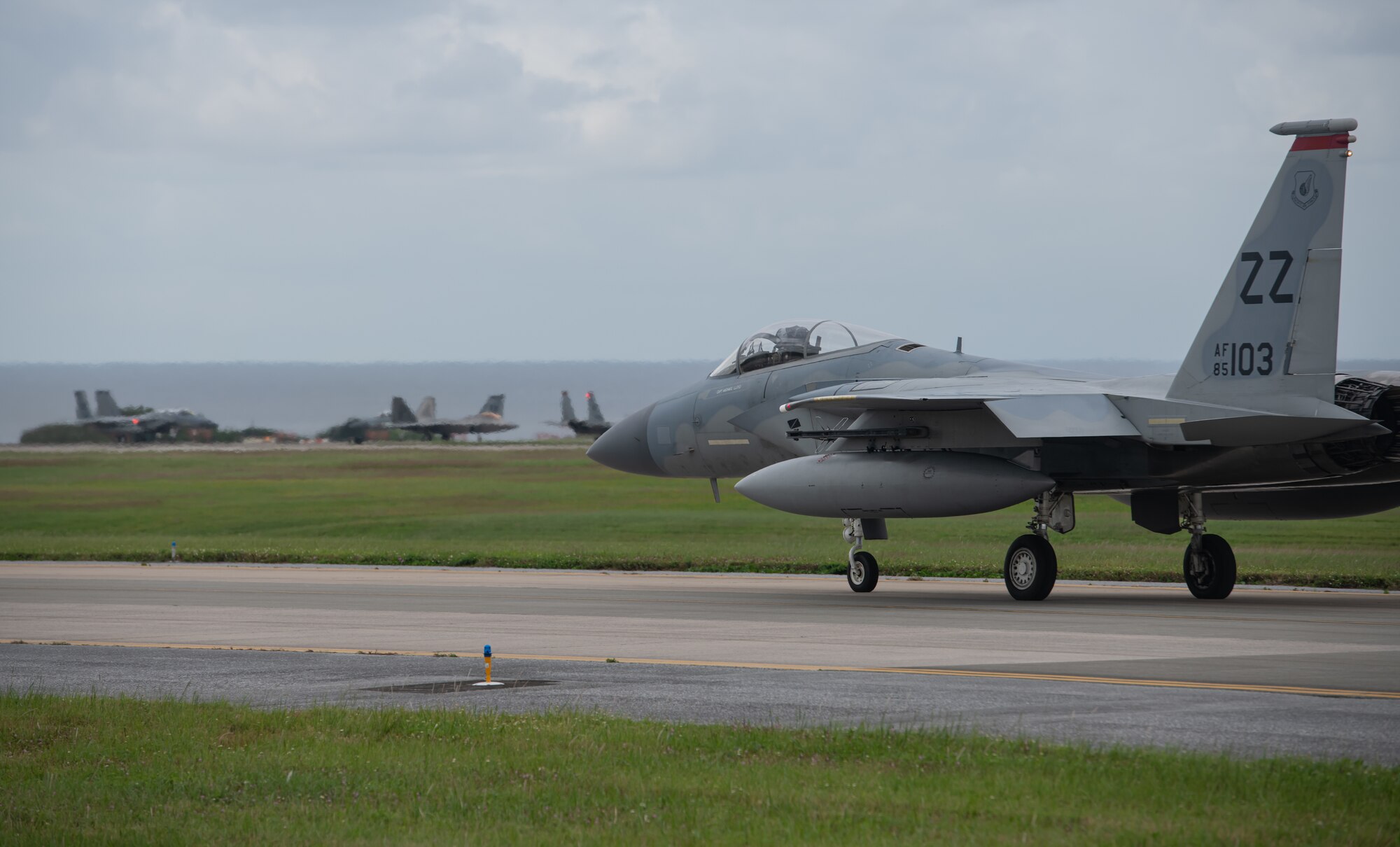 An F-15 Eagle taxis along the runway. In the background there are a mix of F-15s and F-22 Raptors.