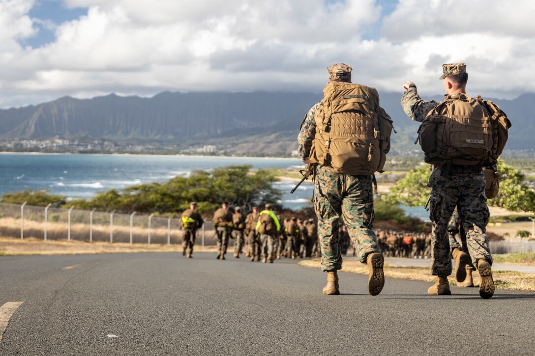 Two Marines walking away from the camera, with a group of Marines far ahead of them on a paved road.