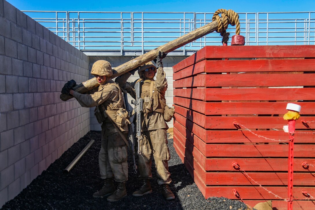 Two Marines carry a pole that hangs over a fence structure and has a rope at one end.