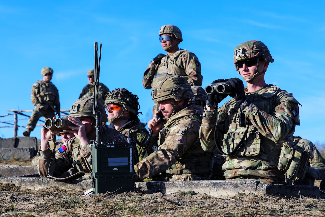 Soldiers look through binoculars while one soldier takes a call during a live fire exercise.