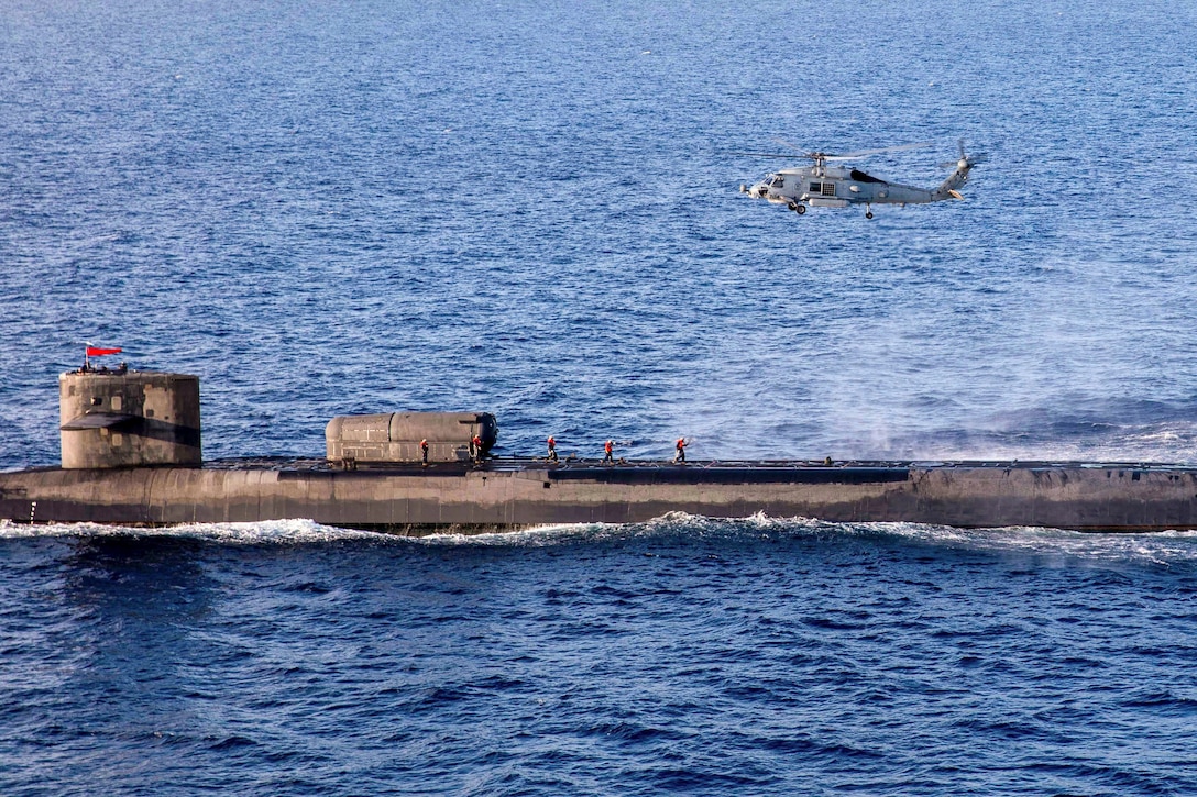 Service members walk on a submarine in a body of water as a helicopter hovers above.