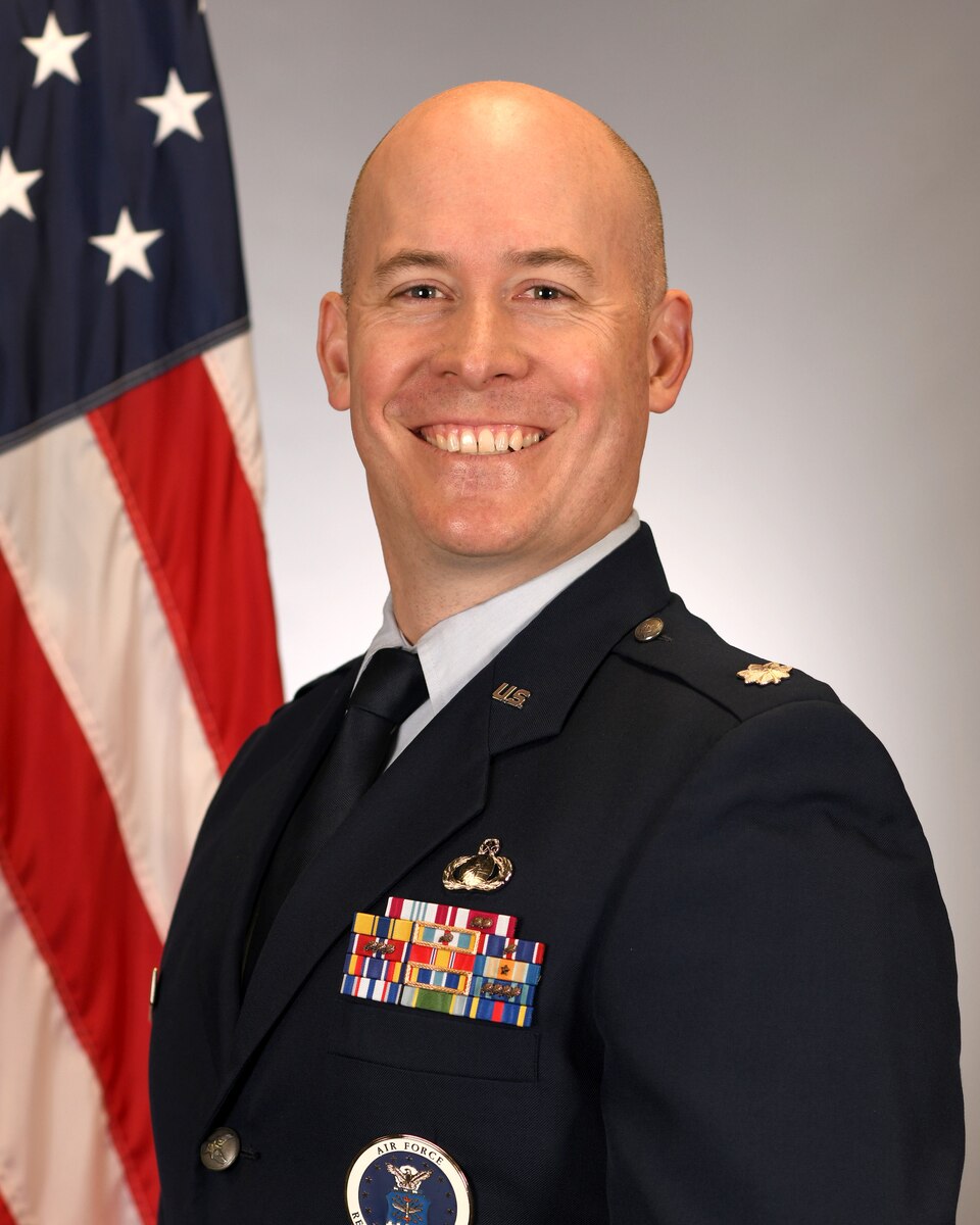 Lt Col Eric Dittman poses for a portrait wearing his Air Force uniform. He is smiling.