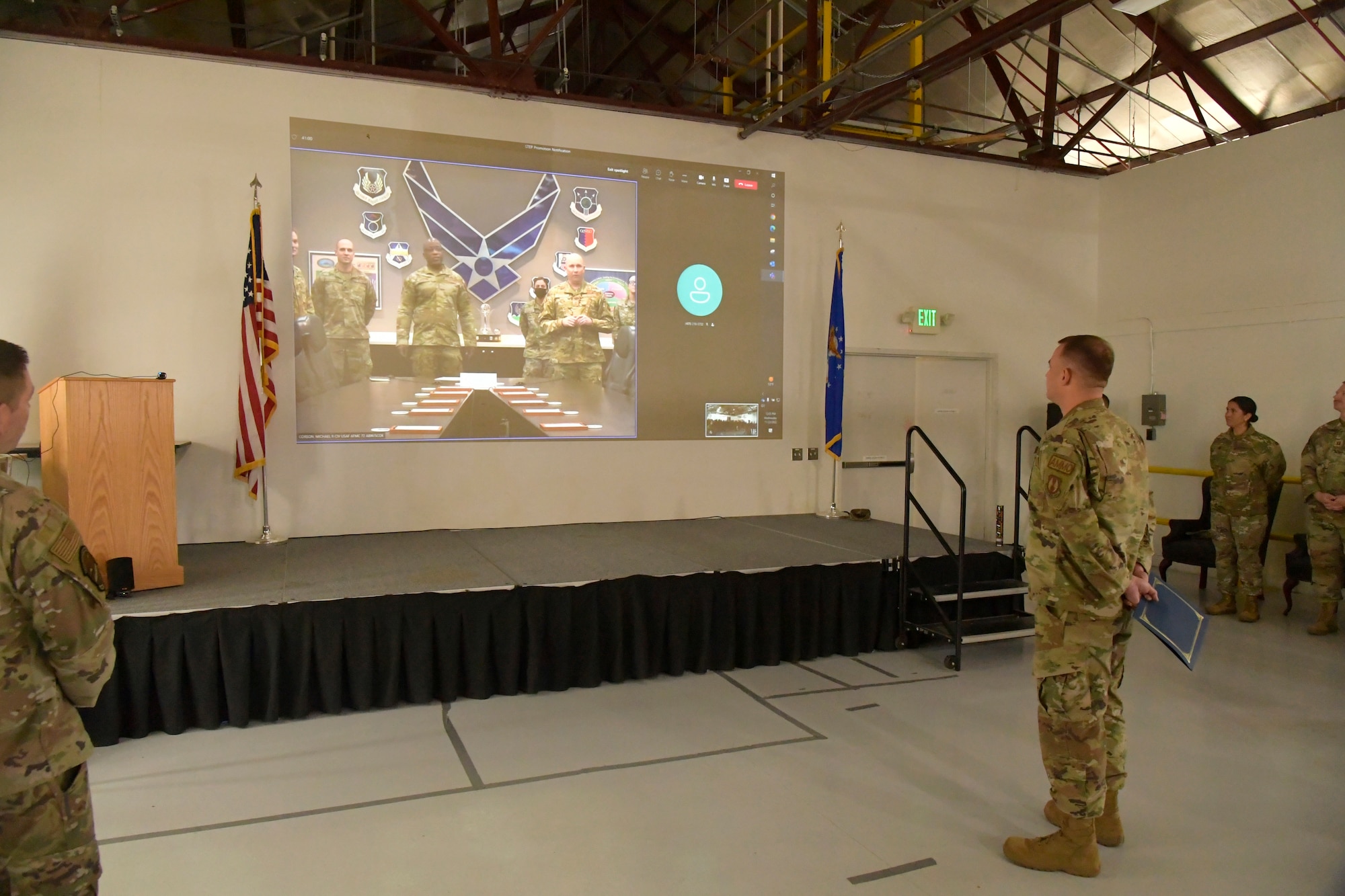 Technical Sgt. Alexander Merrill, 649th Munitions Squadron, stands in front of a screen where Air Force Sustainment Center Commander Lt. Gen. Stacey Hawkins appears congratuating him.