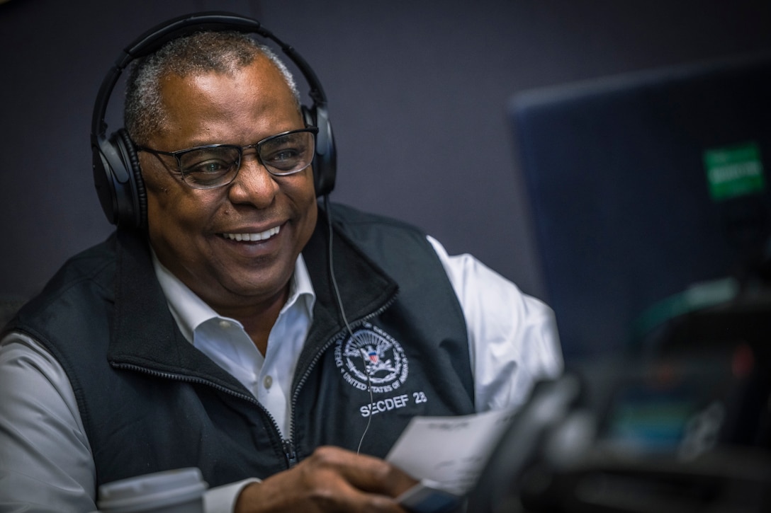 Secretary of Defense Lloyd J. Austin III laughs while wearing headphones and looking at a laptop monitor.