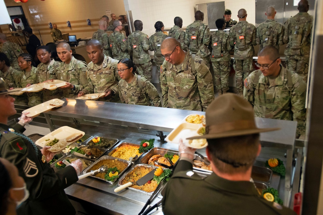 Two rows of trainees stand in a cafeteria; one row faces a buffet being served by two drill sergeants.