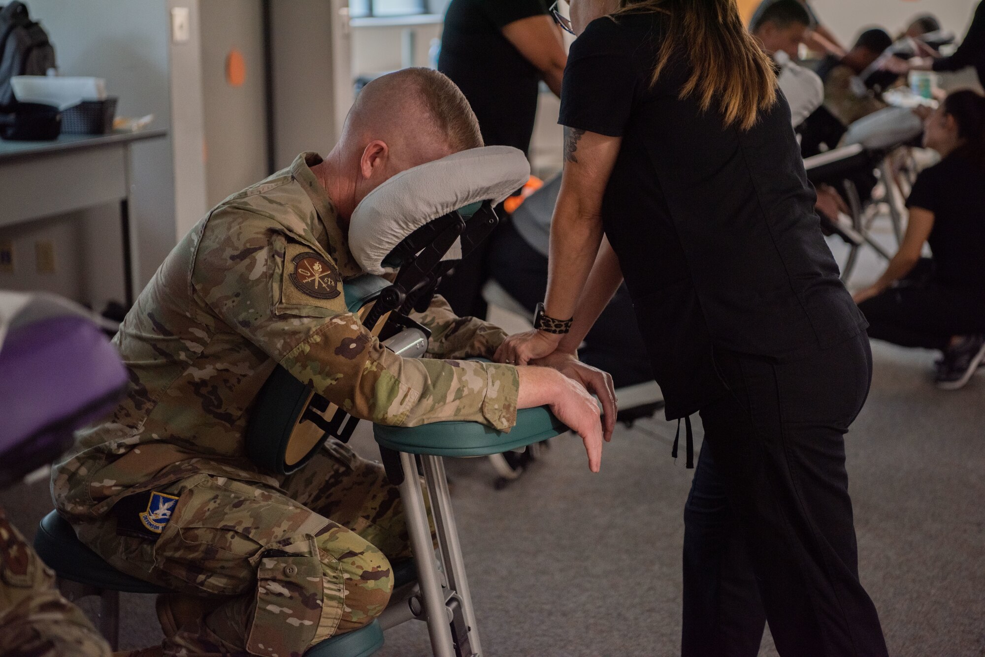 Man in military uniform is sitting down on a massage chair while a massage therapist is massaging him