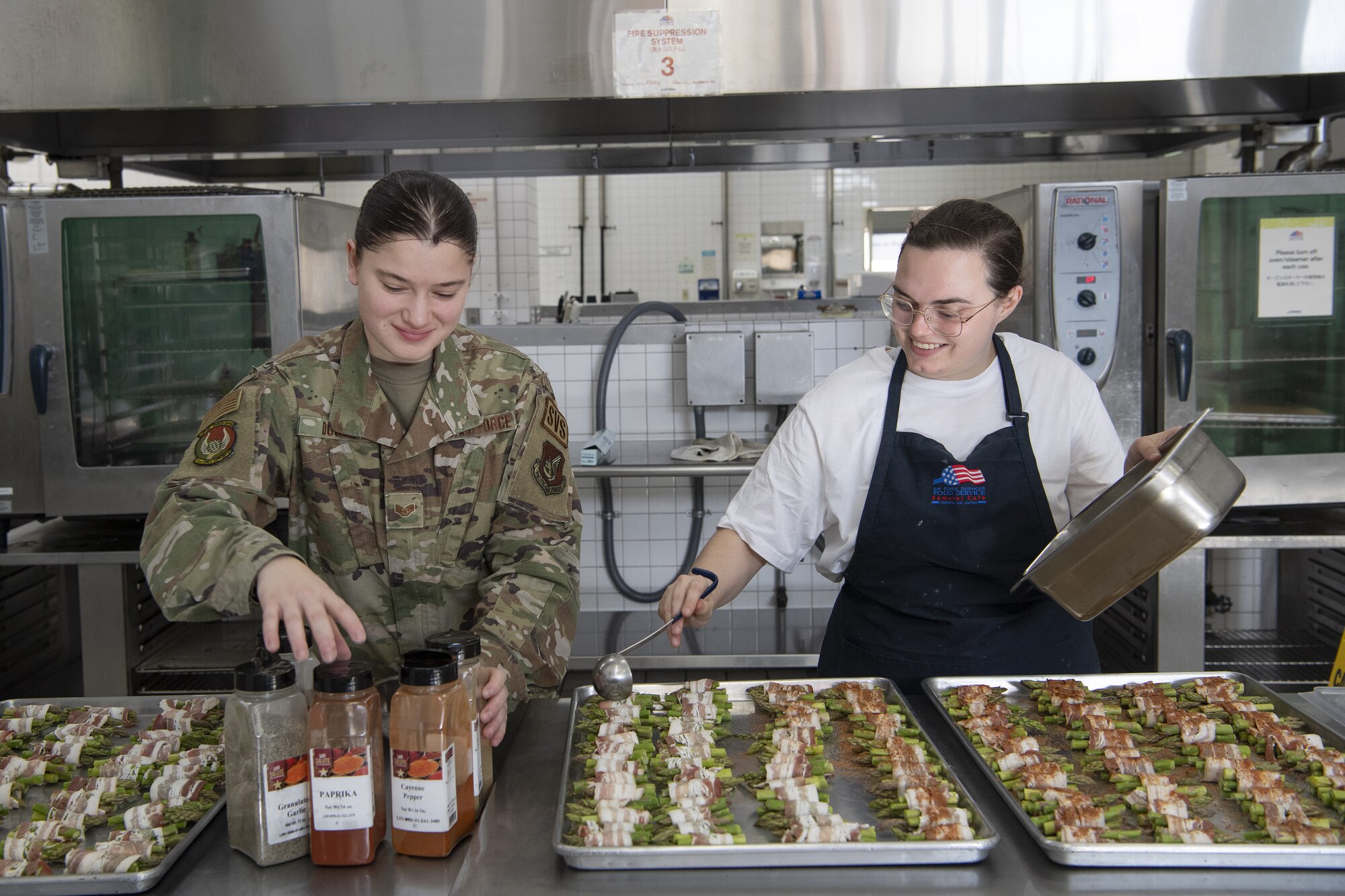 Staff Sgt. Allyson Douty and Staff Sgt. Kylee Masters Thomas, 374th Force Support Squadron food service shift leaders, prepare asparagus wrapped in to serve as part of Thanksgiving meals at the Samurai Café Dining Facility at Yokota Air Base, Japan, Nov. 24, 2022. U.S. Ambassador to Japan Rahm Emanuel, U.S. Air Force Lt. Gen. Ricky Rupp, Commander of U.S. Forces Japan and Fifth Air Force, and 374th Airlift Wing leadership took time to serve and celebrate Thanksgiving Day alongside service members and families assigned to Yokota. (U.S. Air Force photo by Tech. Sgt. Christopher Hubenthal)