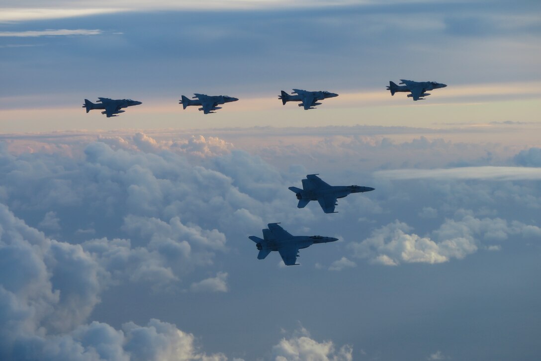 Two U.S. F/A-18E Super Hornets fly below four Italian AV-8B Harrier II aircraft with a cloudy backdrop.