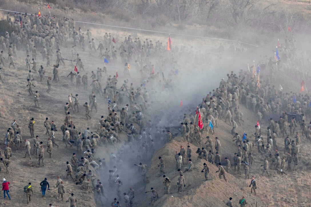 A large group of soldiers is seen from overhead as they tackle a hill with smoke wafting across the center.