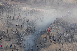 A large group of soldiers is seen from overhead as they tackle a hill with smoke wafting across the center.