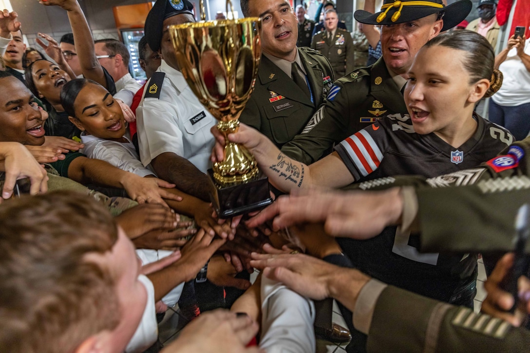 A large group of service members crowd around and reach in to touch a trophy.