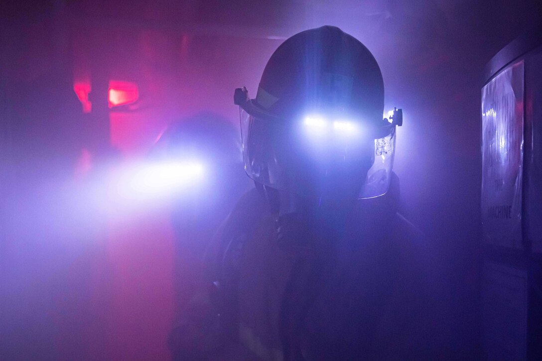 Two sailors wear helmets with headlights and other fire gear during a drill on a ship.