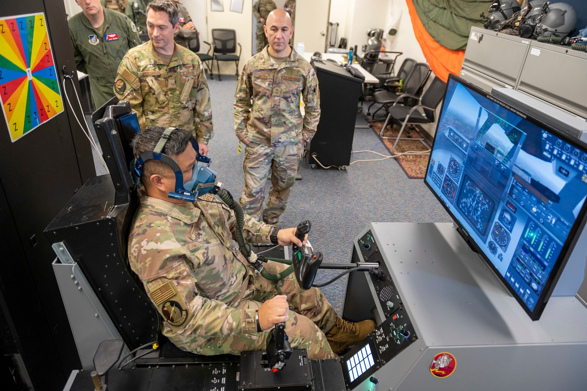 Two soldiers watch a third in a breathing mask try to operate a flight simulator.