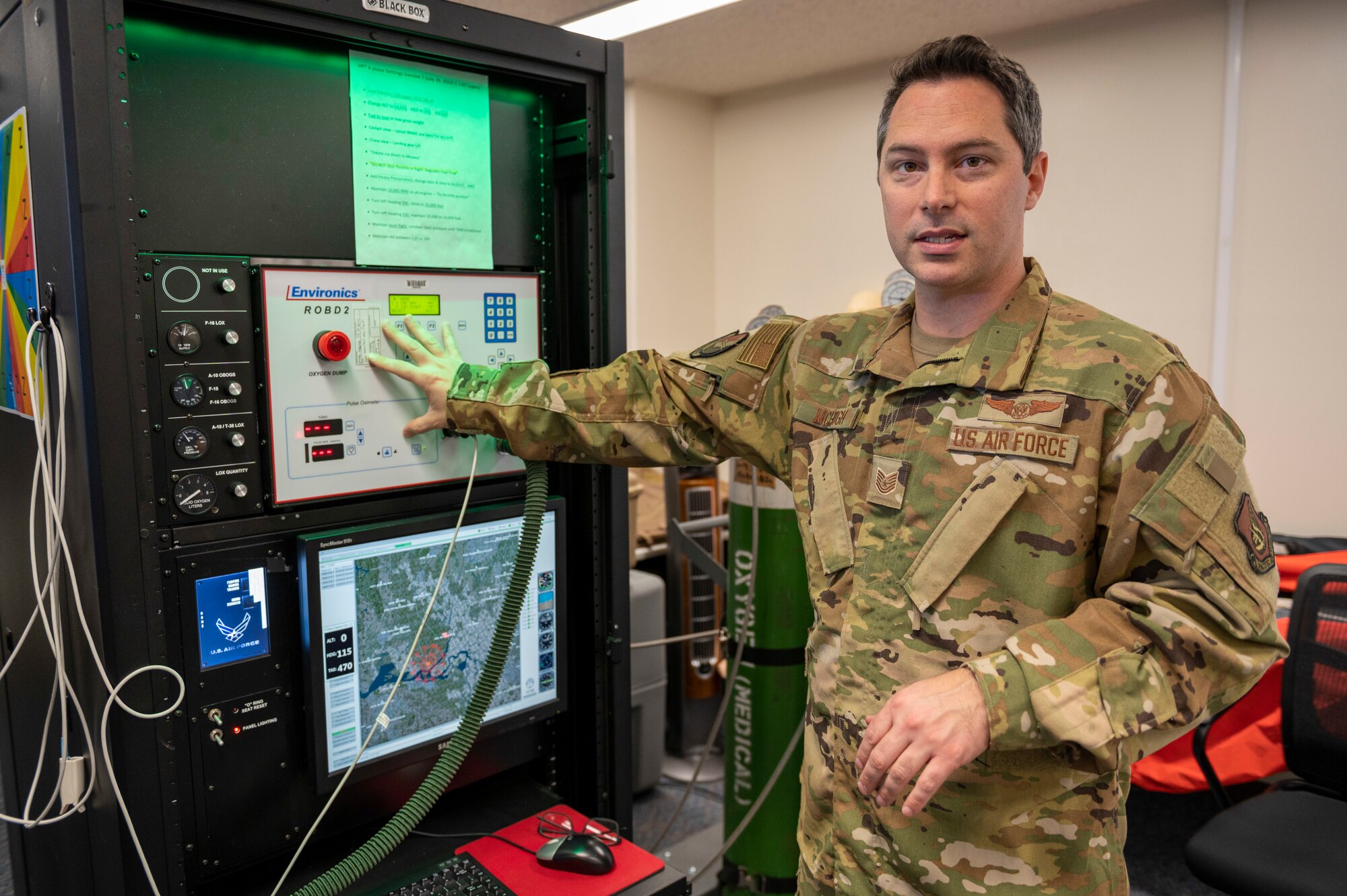 A soldier gestures at a machine hooked up to gas tanks.