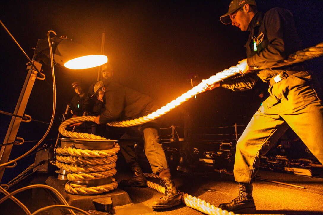 Two sailors handle a line on a ship while others watch.