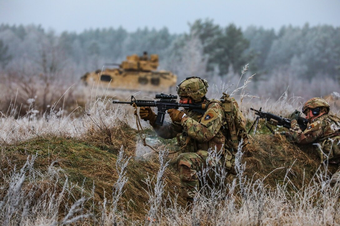 Two soldiers holding weapons and crouching in a field participate in an exercise.