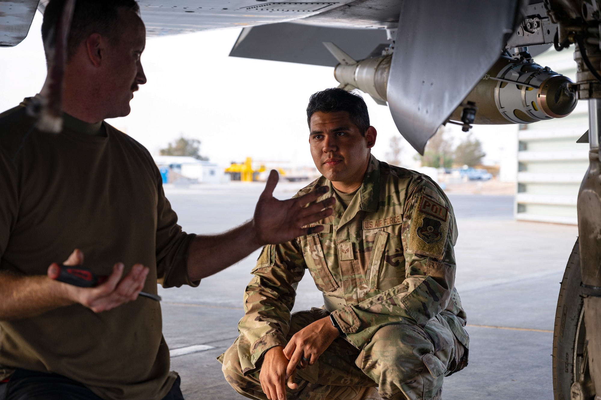 Senior Airman Dennis M. Hatcher, Crew Chief, 389th Expeditionary Fighter Squadron, discusses aspects of his job with Senior Airman Hector J. Gomez, Jr., 332d Expeditionary Logistics Readiness Squadron Fuels Management Flight, as part of the “Maintainer for a Day” program at an undisclosed location, Nov. 10, 2022. Maintainer for a Day is an initiative that pairs Airmen across a variety of career fields with members of aircraft maintenance shops for a hands-on learning experience with F-15E Strike Eagle fighter jets. (U.S. Air Force photo by Tech. Sgt. Richard Mekkri)