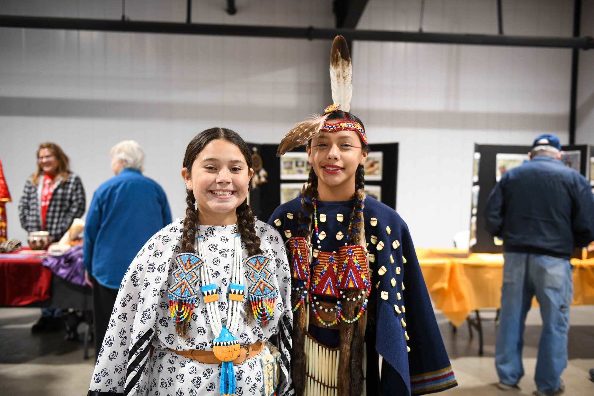 Naima Aiaitson and Bella Portillo, performers with Comanche Nation Youth Dancers, pose for a photo in front of poster boards at the Jackson County Expo Center Nov. 19, 2022 in Altus, Oklahoma. The outfits the dancers wear are all homemade and reflect their heritage with the Comanche Nation. (U.S. Air Force photo by Airman 1st Class Kari Degraffenreed)