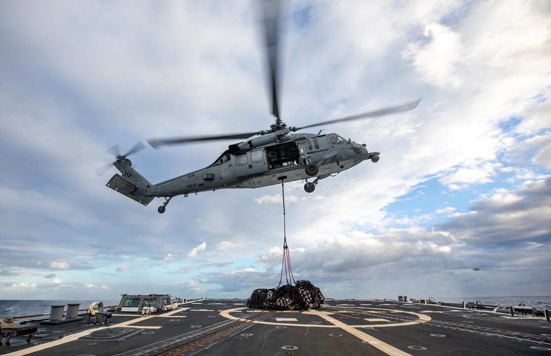 A helicopter lowers a net holding supplies onto a flight deck.