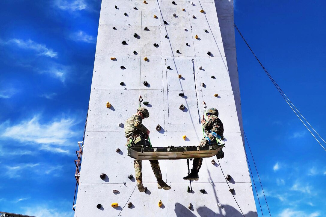 Two soldiers hoist themselves up a tall artificial rock wall.