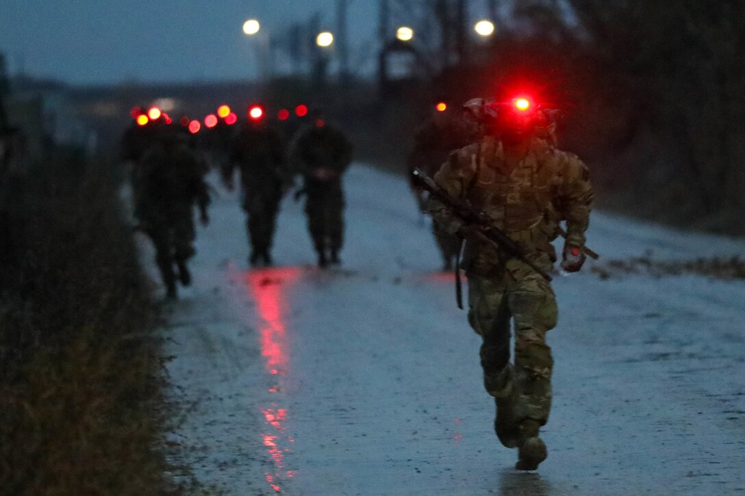 Soldiers wearing headlamps march along a wet road at twilight.