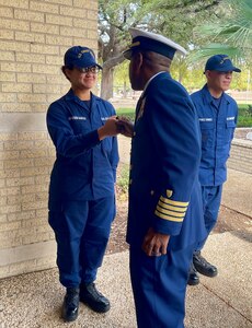 Man, whose back is facing the camera, greets a woman in front of him by doing a fist bump. There is another man on the right side standing. All three people are dressed in a dark blue Coast Guard uniform.