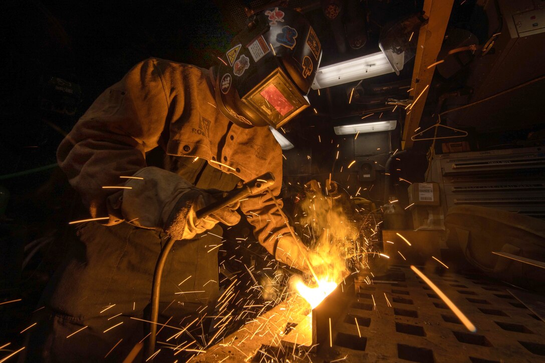 A sailor wearing protective gear welds a piece of metal and sparks fly.
